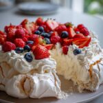 A slice missing from a beautiful Summer Berry Pavlova, topped with whipped cream and a generous mix of strawberries, raspberries, and blueberries.