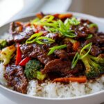 A close-up of a bowl filled with white rice, topped with tender teriyaki beef, broccoli florets, sliced carrots, and red bell peppers, garnished with green onions.