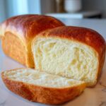A freshly baked loaf of Amish White Bread Recipe, with a slice cut and resting in front.