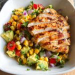 A close-up of a white bowl filled with Avocado Corn Salsa and sliced grilled chicken.
