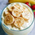 A close-up of Banana Cream Cheesecake Dip in a glass bowl, topped with sliced bananas and brown sugar.