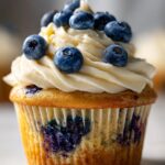 A close-up of a moist blueberry cupcake topped with creamy frosting and fresh blueberries.