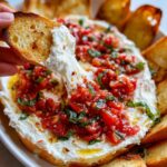 A slice of toasted bread dipping into creamy bruschetta dip, surrounded by more toasted bread.