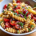 Close-up of a white bowl filled with Bruschetta Pasta Salad, featuring fusilli pasta, cherry tomatoes, red onion, and basil.