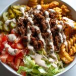 A close-up of a Burger Bowl with Ranch Fries, featuring seasoned ground beef, waffle fries, lettuce, tomatoes, and pickles, drizzled with ranch dressing.