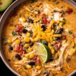 A close-up of a steaming bowl of Creamy Chicken Enchilada Soup Recipe, topped with shredded cheese, black beans, corn, and a lime wedge.