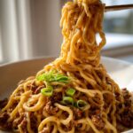 Close-up of creamy garlic ramen with ground beef, garnished with green onions, being lifted by chopsticks.