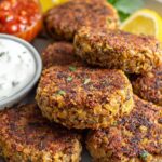 A stack of golden-brown Crispy Lentil Patties served with a side of dipping sauce and lemon wedges.