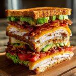 A tall, triple-decker Crispy Ultimate Club Sandwich with turkey, bacon, cheese, avocado, tomato, and lettuce on toasted bread.