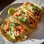 Three delicious shrimp tacos with avocado, shredded cabbage, cilantro, and a creamy sauce, served with a lime wedge. Easy Shrimp Tacos Recipe 5-Minute.