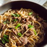 Close-up of a bowl filled with Garlic Beef Noodle Bowls Recipe, featuring tender beef strips, noodles, and green onions.