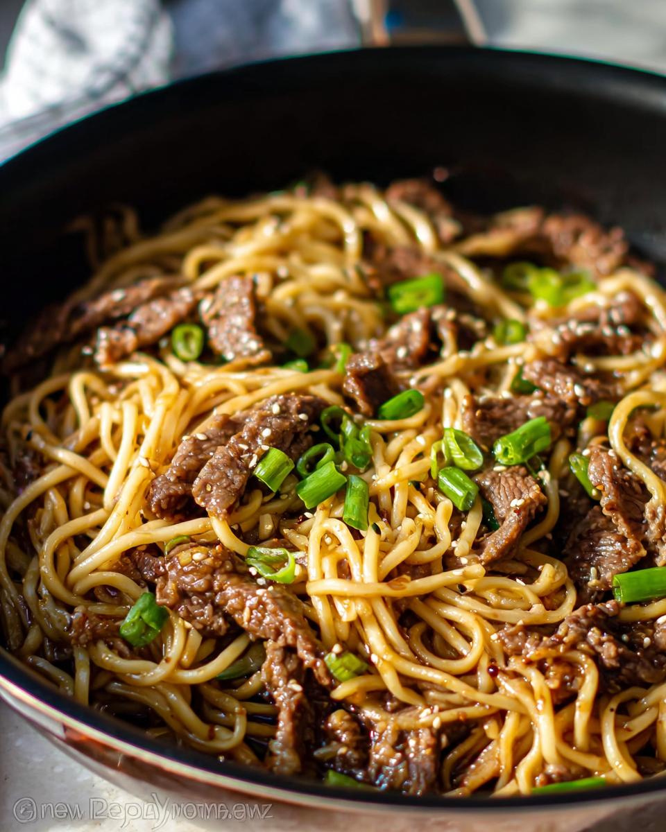 Speedy Garlic Beef Noodle Bowls Recipe 5 Close-up of a pan filled with Garlic Beef Noodle Bowls, featuring tender beef strips, noodles, and green onions.