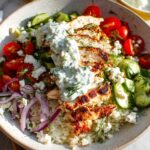 A close-up of Greek Chicken Bowls with sliced grilled chicken, rice, cucumber, tomatoes, feta, red onion, and creamy tzatziki sauce.