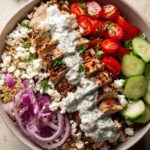 A close-up of a Greek Chicken Power Bowl featuring grilled chicken, rice, tomatoes, cucumbers, feta, red onion, and tzatziki sauce.