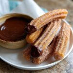 A close-up of freshly made homemade churros coated in cinnamon sugar, served with a bowl of rich chocolate sauce.