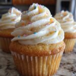 Close-up of three fluffy homemade vanilla cupcakes topped with white frosting and colorful sprinkles.