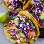 Close-up of three Honey Lime Salmon Tacos filled with flaky salmon, avocado, red cabbage, and crumbled cheese.