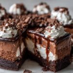 A decadent slice of Hot Fudge Sundae Brownie Cheesecake with layers of chocolate mousse, cheesecake, and brownie base, topped with whipped cream and hot fudge.