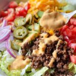 Close-up of a Keto Burger Bowl with seasoned ground beef, lettuce, tomatoes, avocado, jalapeños, red onion, cheese, and drizzled with easy burger sauce.