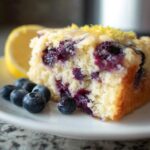 A slice of Lemon Blueberry Breakfast Cake topped with glaze and lemon zest, served with fresh blueberries and a lemon slice.