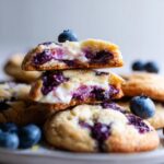 A stack of delicious lemon blueberry cheesecake cookies, with a bite taken out of the top one to reveal the creamy cheesecake filling and blueberries.