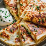 Close-up of golden-brown Mac and Cheese Quesadilla slices, oozing with melted cheese and sprinkled with chives, served with sour cream.