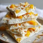 A stack of Easy Mango Coconut Frozen Yogurt Bark pieces, topped with mango chunks, coconut flakes, and slivered almonds.