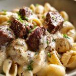 Close-up of tender steak bites served over creamy Alfredo shell pasta, garnished with parsley.