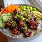 A close-up of a Sticky Asian Chicken Poke Bowl Easy Rice Bowl with glazed chicken, rice, avocado, cucumber, edamame, and shredded carrots.