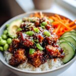 Close-up of a delicious Sticky Sesame Chicken Poke Bowl with rice, edamame, avocado, cucumber, and shredded carrots.