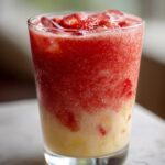 A tall glass filled with a vibrant Strawberry Colada Smoothie, layered with red strawberry and creamy yellow smoothie, topped with fresh strawberries and ice.
