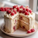 A slice of White Chocolate Raspberry Cake with Cream Cheese Frosting, topped with fresh raspberries and white chocolate chips.